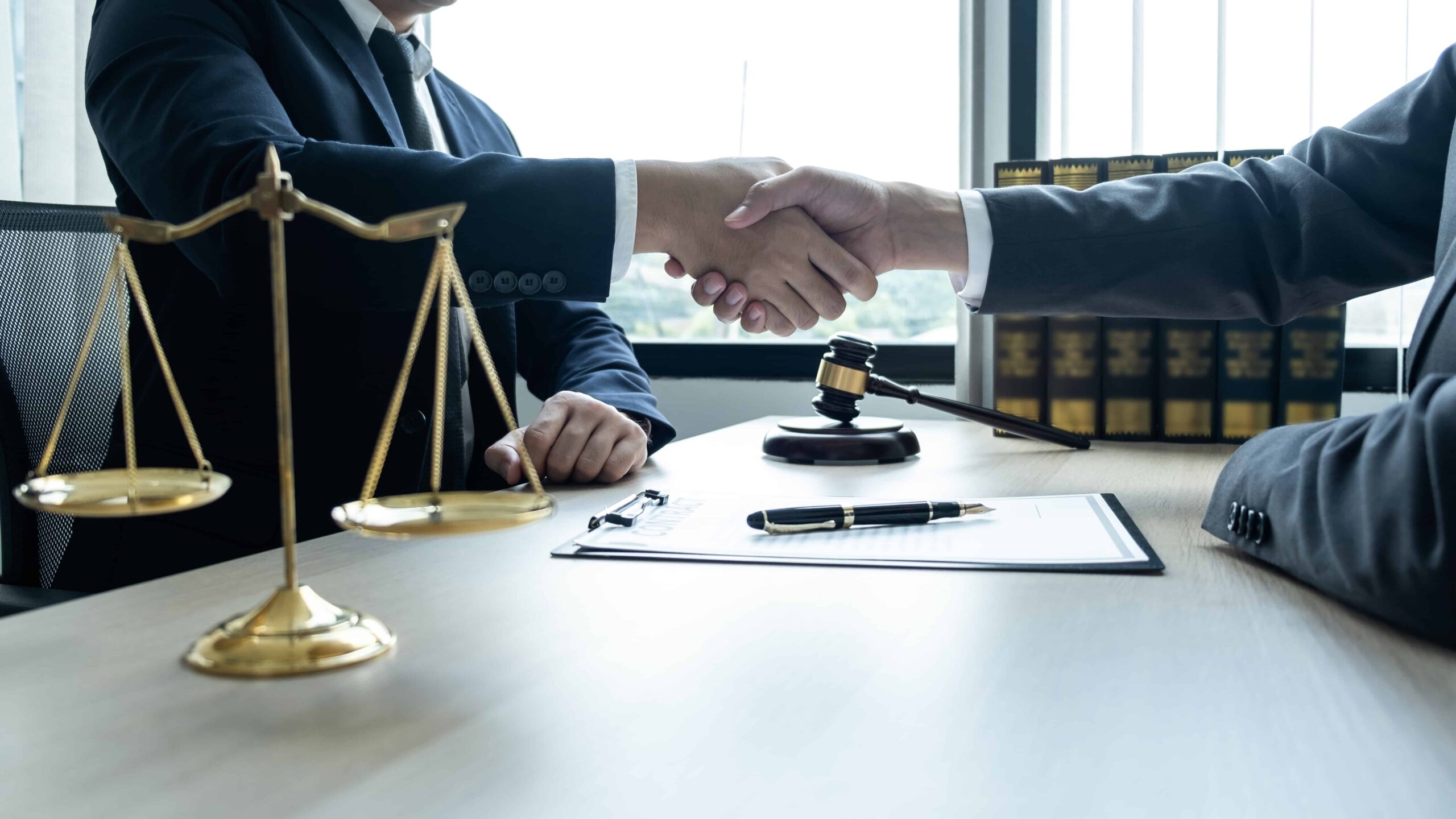 Attorneys shaking hands across a desk with scales of justice, gavel, and legal documents, symbolizing a legal settlement or agreement